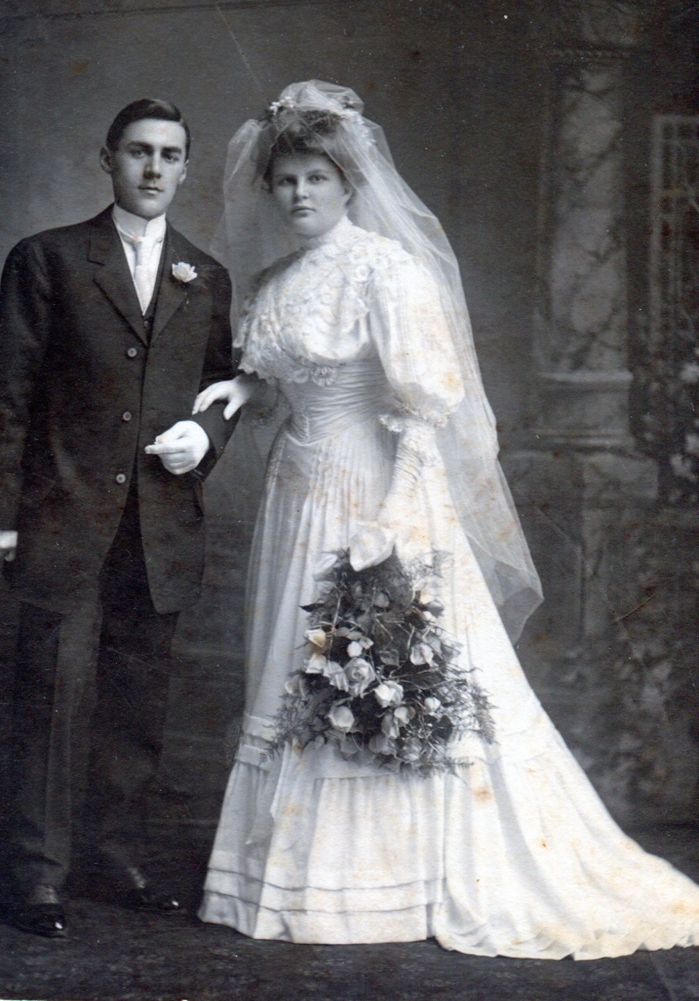 Man and bride wearing a white wedding gown with a high neckline, puffy bodice, and long flowing skirt. Dress is decorated by lace ruffles.