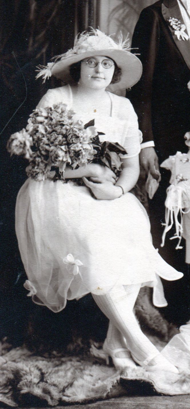 Bridesmaid on left with large hat, chiffon dress with square neckline and slightly gathered skirt and bouquet of long stemmed flowers.