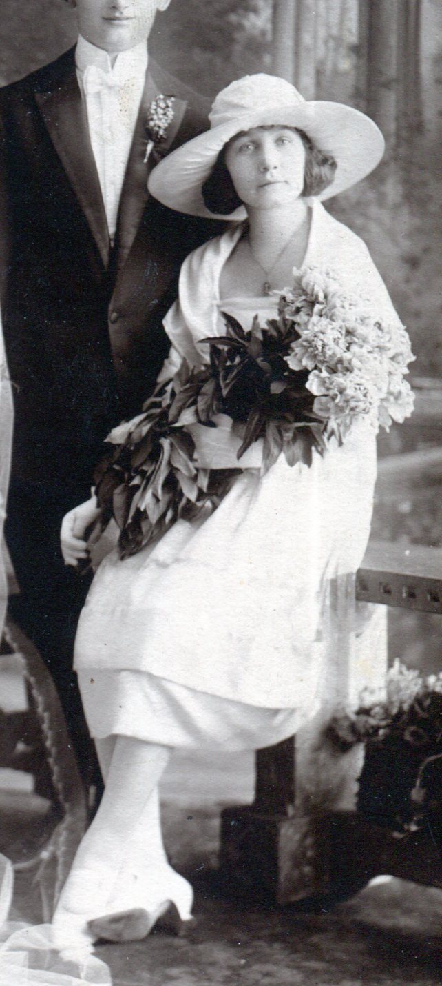 Bridesmaid on right with large hat and dress with square neckline, 2 layer skirt, shawl and bouquet of long stemmed flowers.