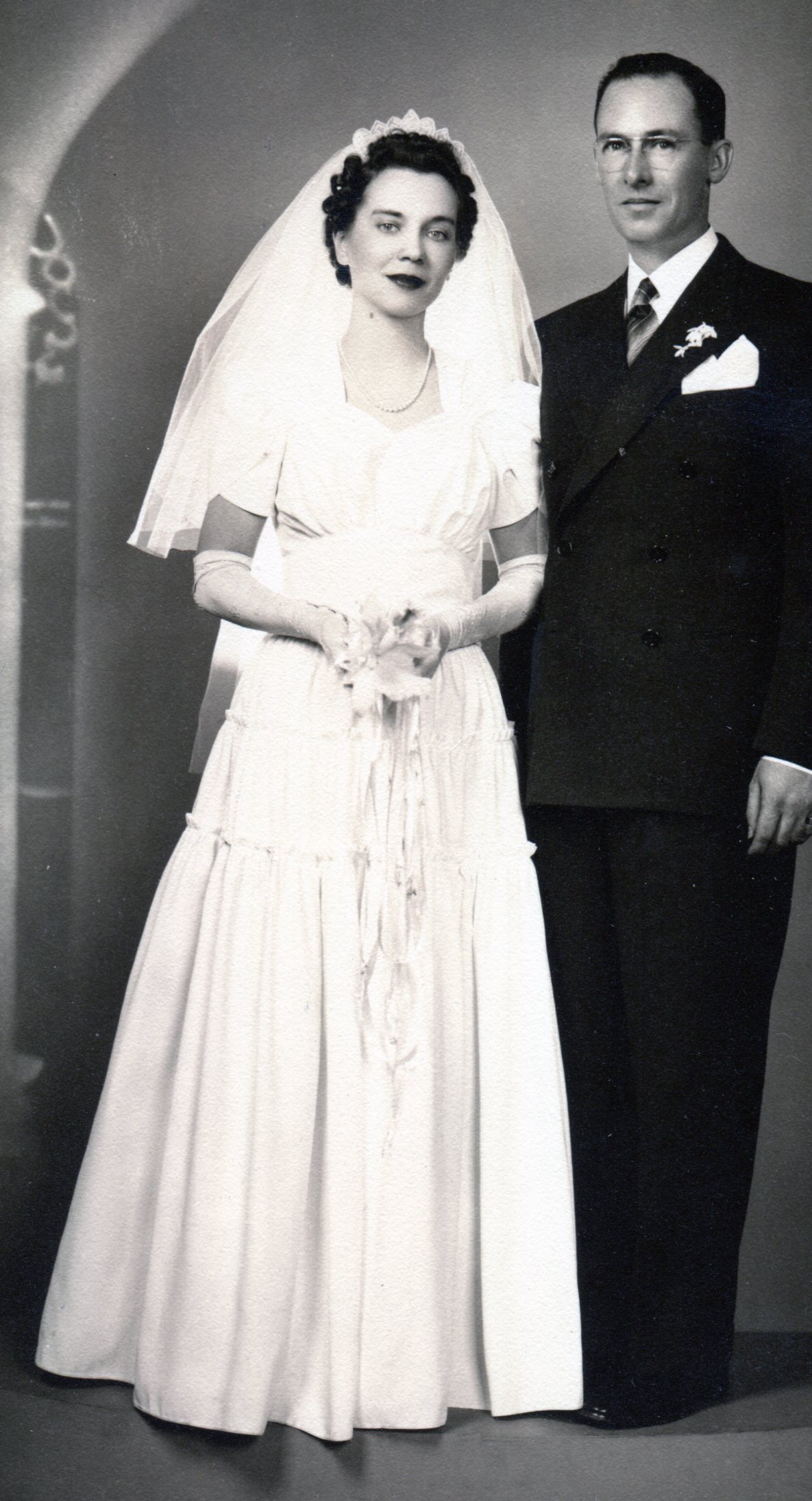 1941 Bride wearing a white taffeta gown with gathered bust line above a flat stomacher, gathered skirt in 3 gathered tiers, and long glove with the groom