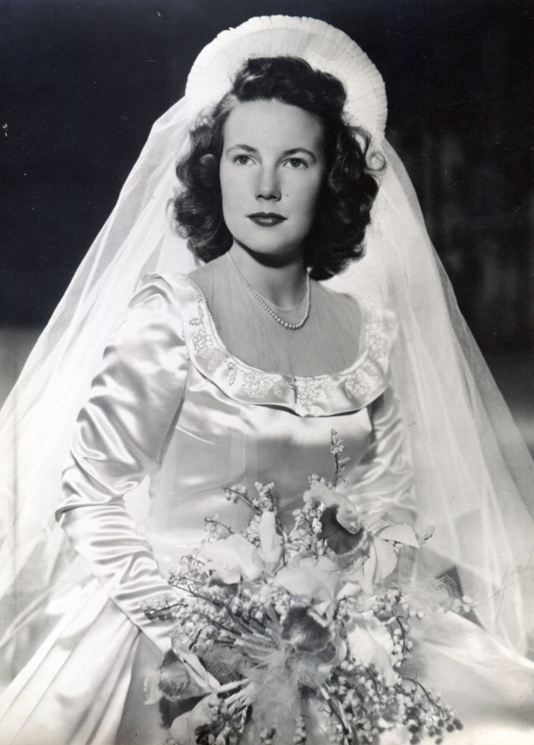 Bride in full skirted white satin dress with a scoop neckline with a ruffle, neckline filled in by sheer fabric and ruffled headdress sticking up above her face.