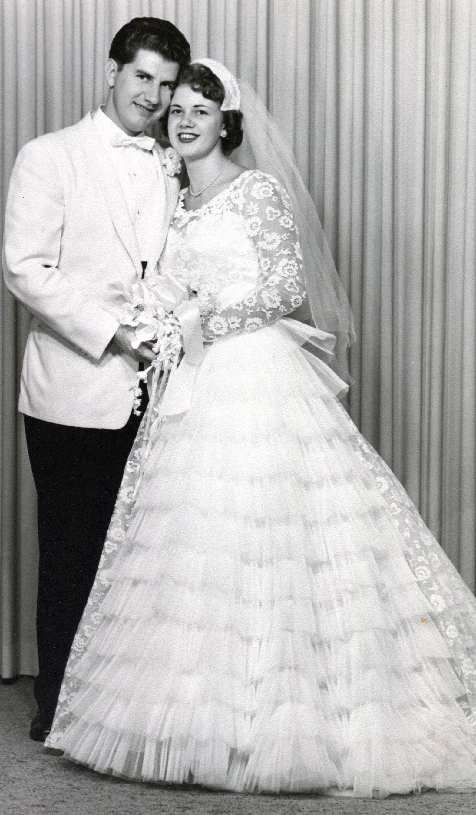 Bride in white lace dress with full skirt made of many ruffles. Groom wears white jacket.