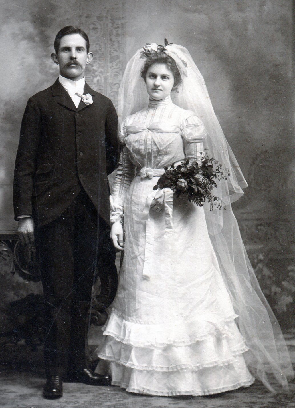 Bride with high neck white dress with bands of flat lace on the bodice, long sleeves, and full length skirt with three rows of ruffles at the bottom.