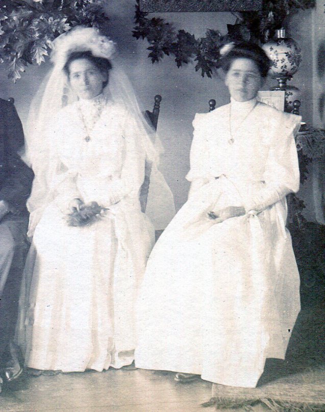 Bride and Maid-of-Honor both wearing high neck dresses with long sleeves and long skirts sitting on chairs next to each other.