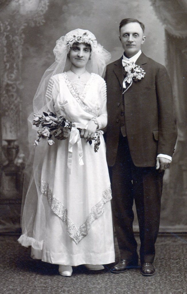 1914 Bride in white dress with overlapping pieces with flat embroidered band trim and Groom with large bouttonniere. 