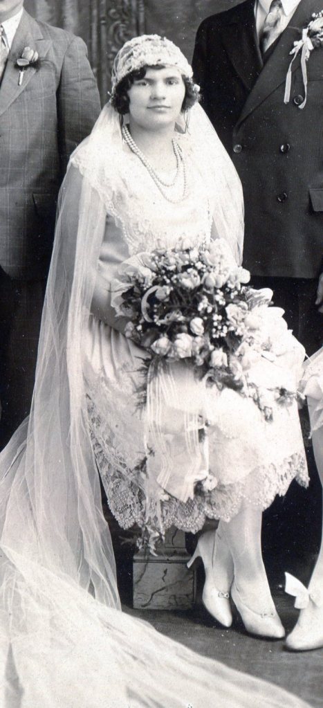 The 1925 Bride sitting down holding her bouquet and wearing a sleeveless bridal gown with a lace fabric covering part of the solid underneath fabric.
