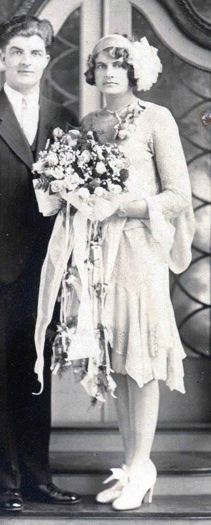 Brides maid in dress of sheet lace fabric over a solid base, standing holding the bridal bouquet. Shows the pointed handkerchief hem of her dress.