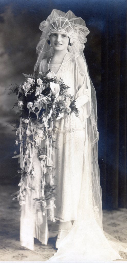 1923 Bride in elegant straight light fabric with white beaded pattern on gown and wearing a large headpiece with puffy netting and a pearl bead crown.
