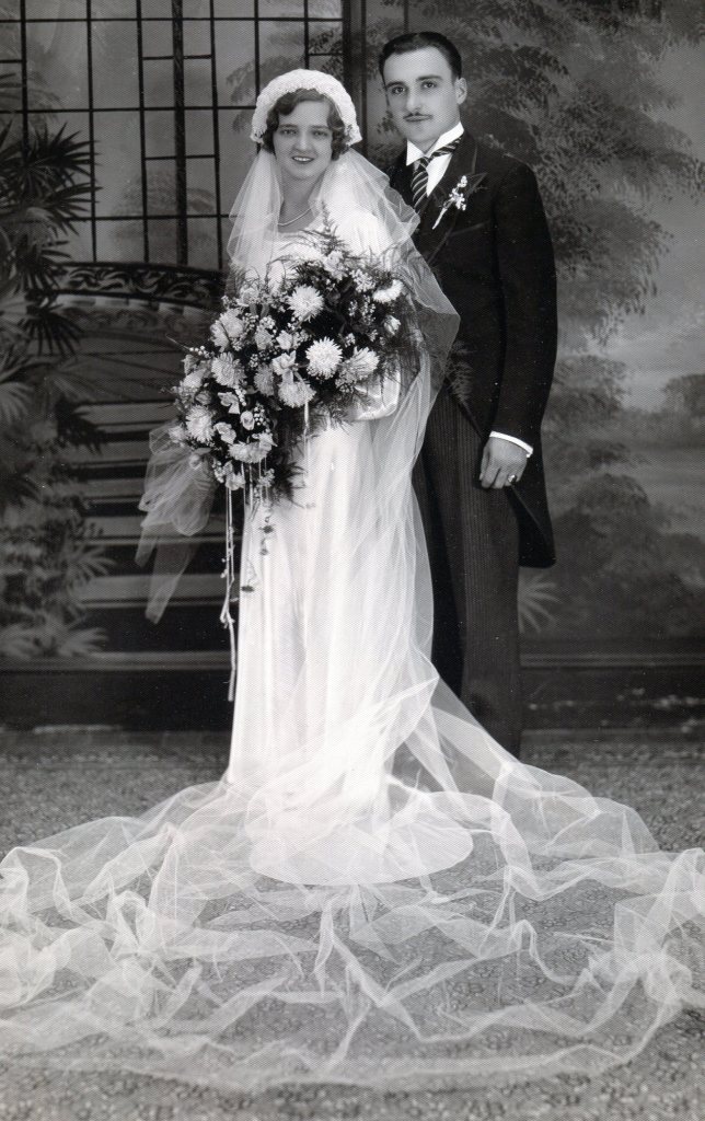 1930s Bride and Groom. The bride wears a long white satin dress and long veil. The groom wears a black cut-away coat with tails.