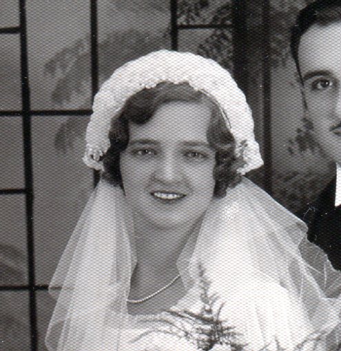 Bride wears a headpiece of stiffened fabric covered with pearls and white net veil attached at back.