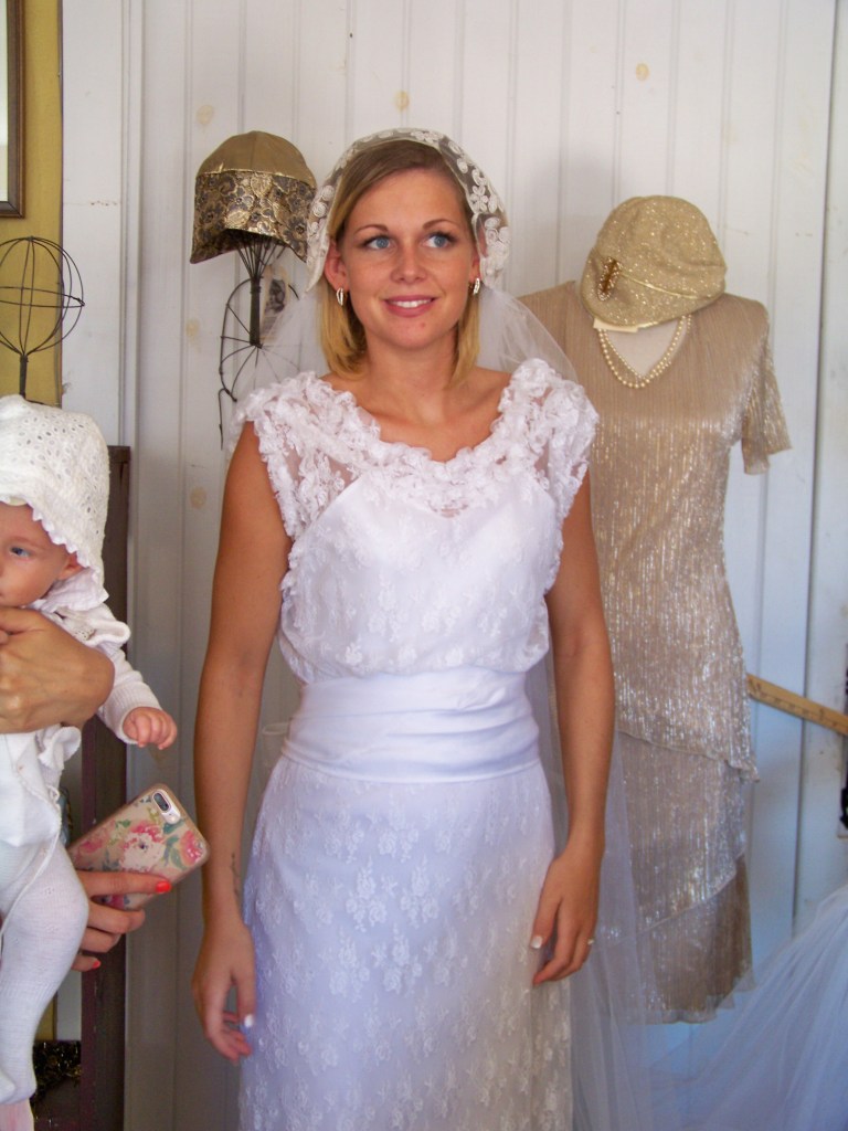 1930s bride in sleeveless white sheet lace dress with V-neckline with small lace ruffles and wide white cumberbund.
