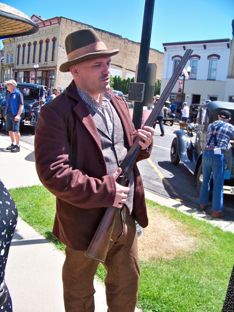 Man in brown suit jacket and brown fedora holding shotgun.