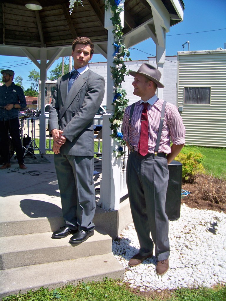 Groom in grey double breasted suit and Best Man in grey pants with suspenders, red and white striped shirt with white collar and red tie and grey fedora.