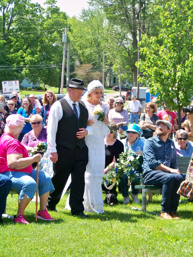 Father of the Bride walks bride down aisle with car show spectators watching.