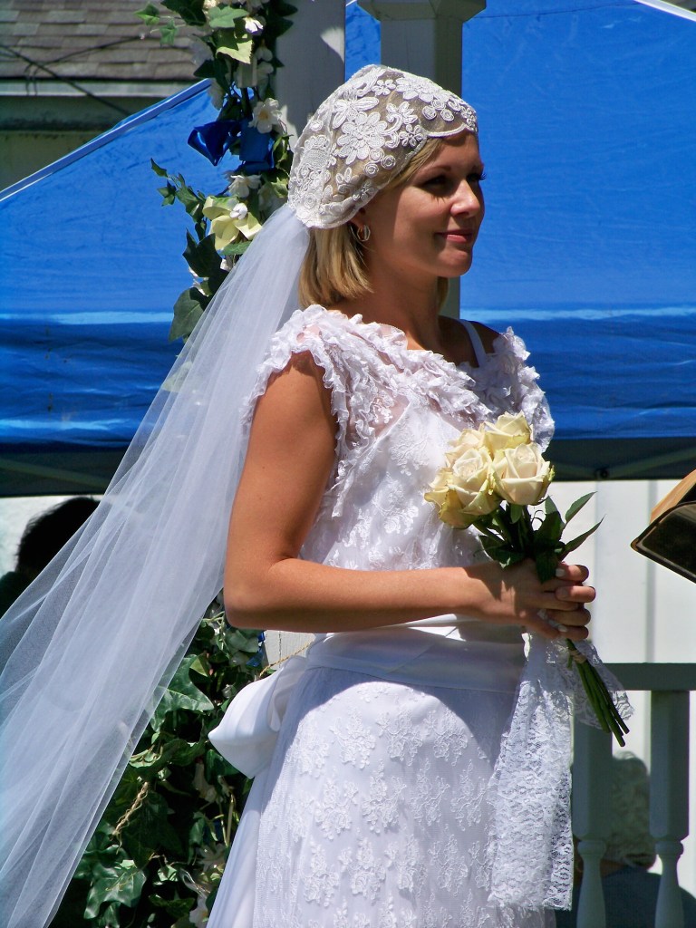 Bride standing in long white dress, bridal cap with long veil, and holding four yellow long-stemmed roses. 