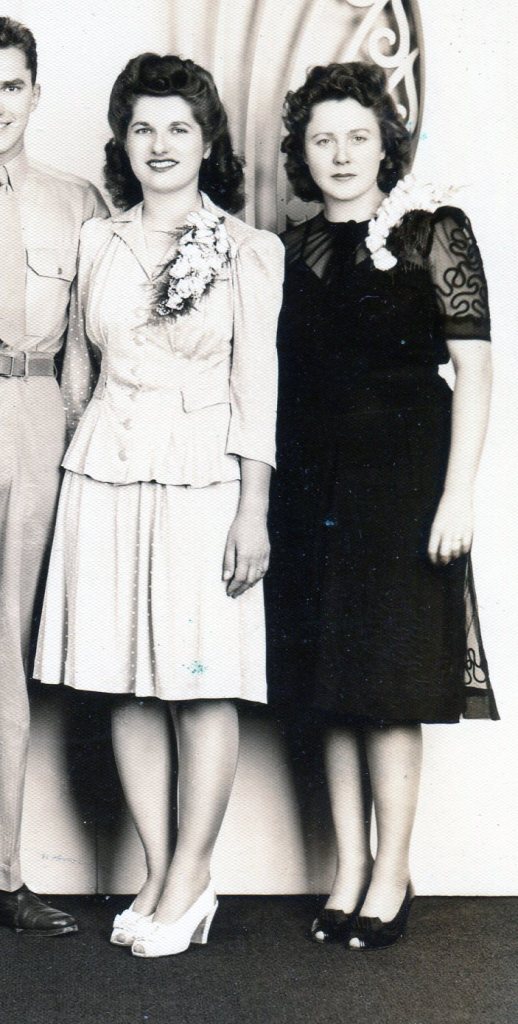 Maid of Honor wearing a black party dress of decorated sheer fabric over a solid black fabric wears a large corsage and stands next to bride.