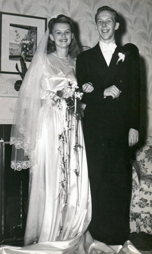 A smiling Bride and Groom. The Bride wears a long white satin dress, a net veil, and small bouquet of flowers. The Groom is in a black suit.